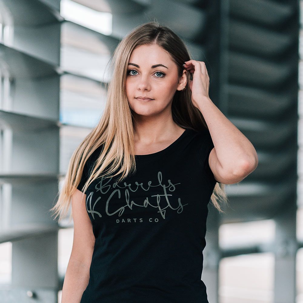Barrels and Shafts dames-T-shirt - zwart Eine Frau trägt ein schwarzes T-Shirt mit der Aufschrift „Barrels and Shafts Darts Co“. Sie steht vor einem modernen, lichtdurchfluteten Hintergrund.