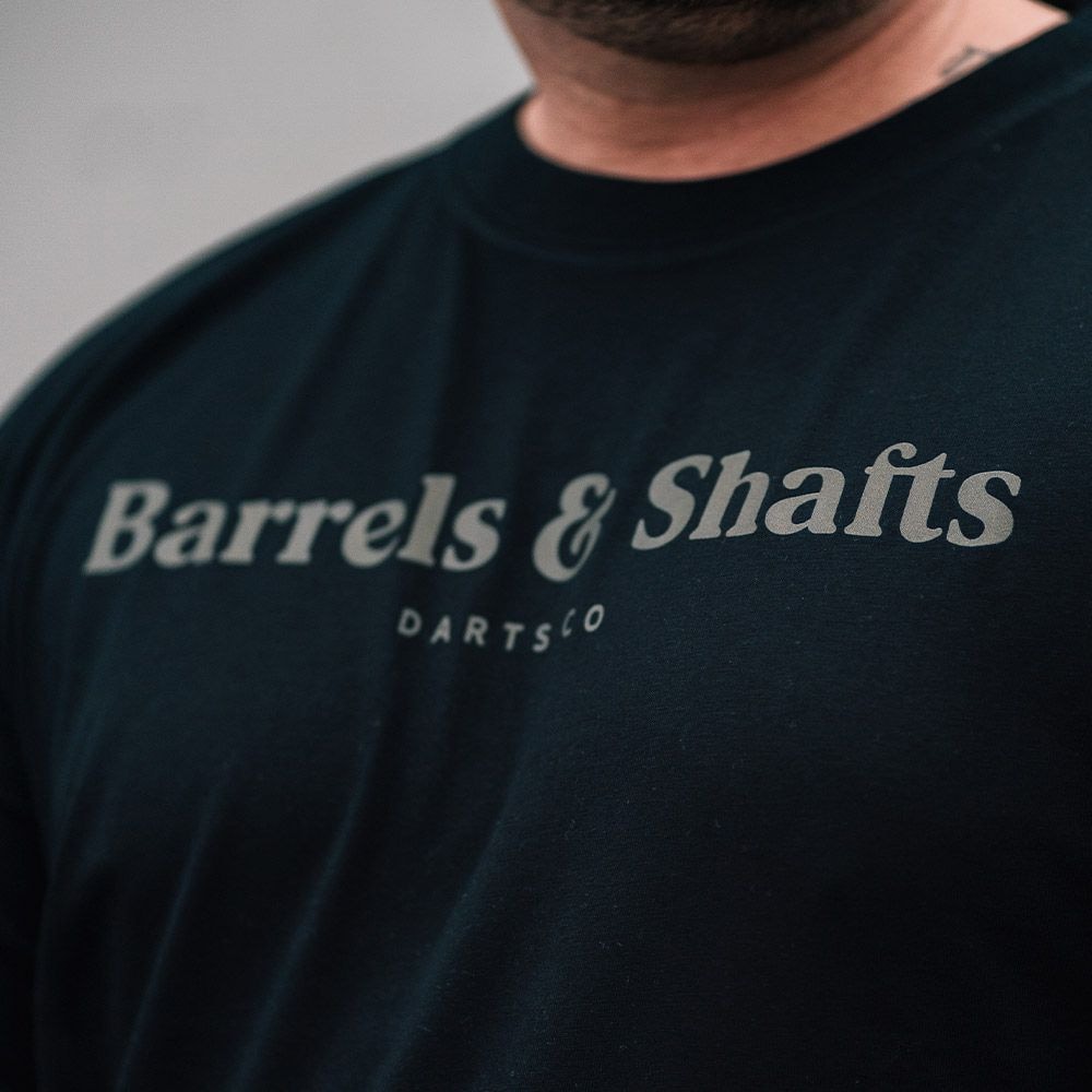 Vaten en schachten T-shirt - zwart Das Bild zeigt ein schwarzes T-Shirt mit der Aufschrift "Barrels & Shafts". Darunter steht in kleineren Buchstaben "DARTSCO".