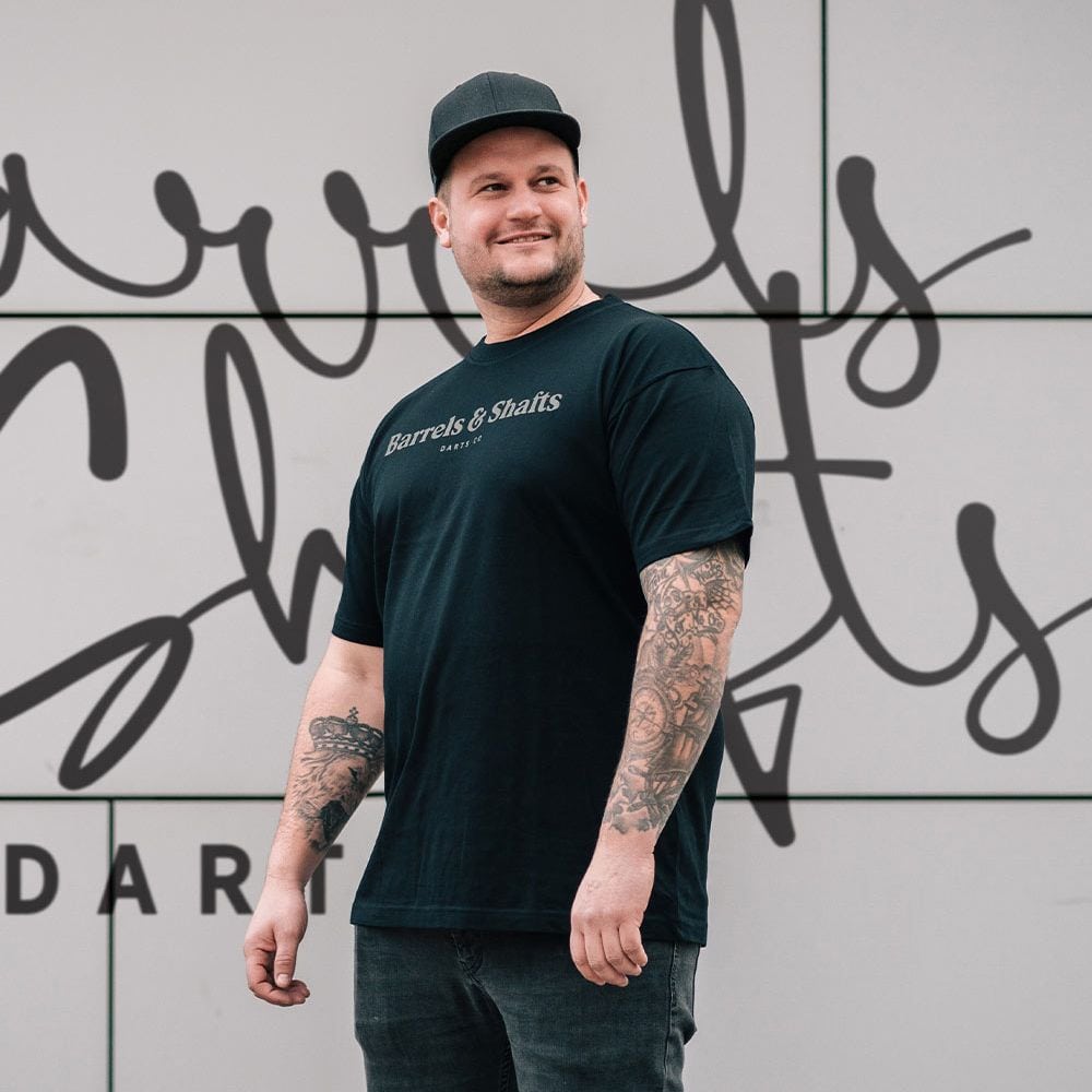 Vaten en schachten T-shirt - zwart Ein Mann trägt ein schwarzes T-Shirt mit der Aufschrift „Barrels & Shafts“. Er steht vor einer Wand mit passender Schrift im Hintergrund.