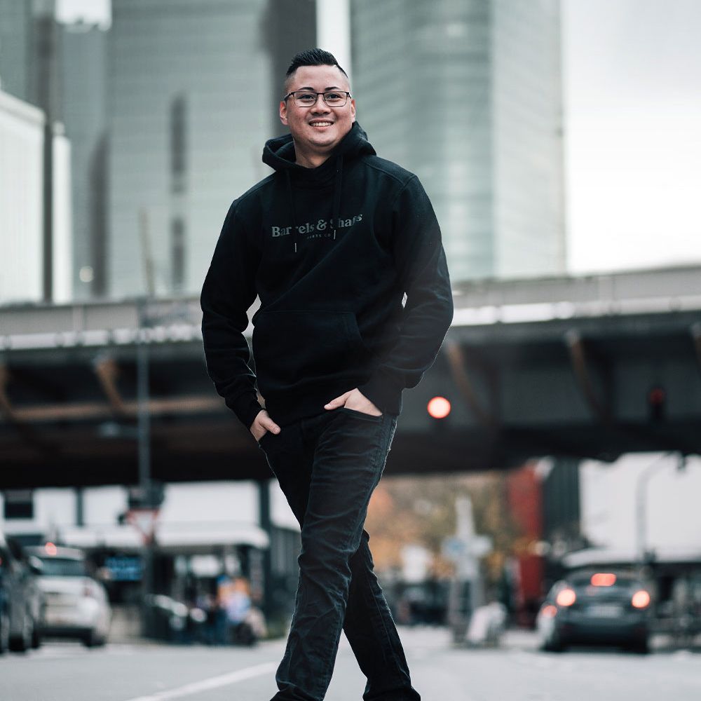 Vaten en schachten hoodie - zwart Eine Person trägt einen schwarzen "Barrels and Shafts" Hoodie und steht entspannt auf einer Straße. Im Hintergrund sind unscharfe Gebäude und Autos zu sehen.