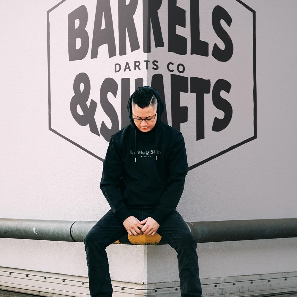 Vaten en schachten hoodie - zwart Eine Person trägt einen schwarzen "Barrels and Shafts" Hoodie und sitzt auf einer Bank. Im Hintergrund ist ein großes Wandlogo mit der Aufschrift "Barrels & Shafts Darts Co" zu sehen.