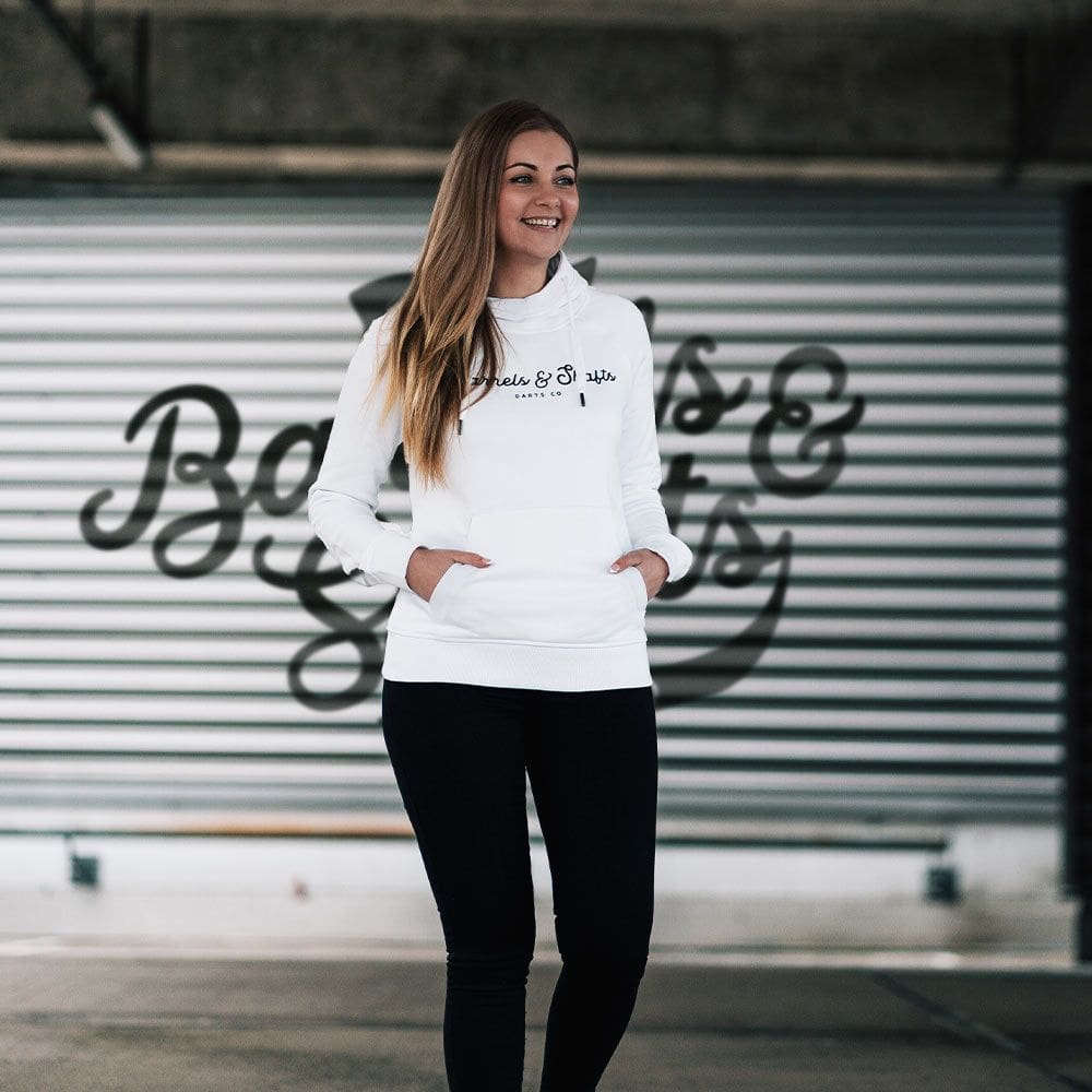 Barrels and Shafts Dameshoodie - Wit Eine Frau trägt einen weißen "Barrels and Shafts" Damen Hoodie. Sie steht vor einem Hintergrund mit großem Schriftzug und lächelt.