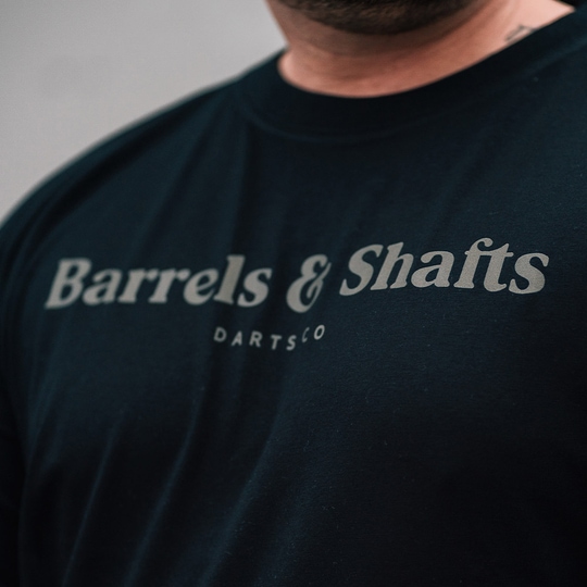 Vaten en schachten T-shirt - zwart Das Bild zeigt ein schwarzes T-Shirt mit der Aufschrift "Barrels & Shafts". Darunter steht in kleineren Buchstaben "DARTSCO".