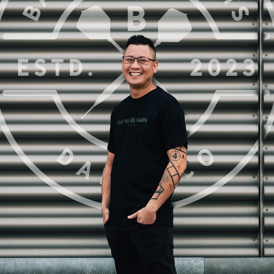 Vaten en schachten T-shirt - zwart Ein Mann trägt ein schwarzes "Barrels and Shafts"-T-Shirt und steht vor einer metallenen Wand mit großem Logo. Er lächelt und hat markante Tattoos am rechten Arm.