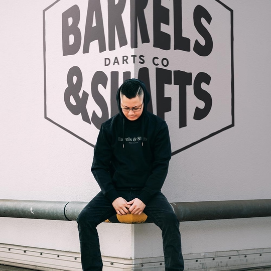 Vaten en schachten hoodie - zwart Eine Person trägt einen schwarzen "Barrels and Shafts" Hoodie und sitzt auf einer Bank. Im Hintergrund ist ein großes Wandlogo mit der Aufschrift "Barrels & Shafts Darts Co" zu sehen.