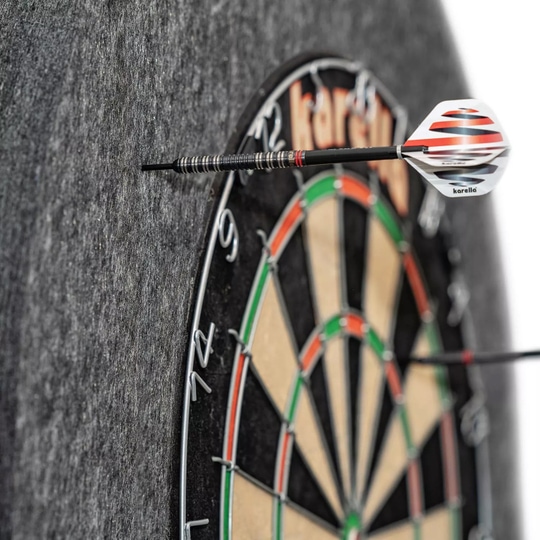 Karella Ultimo stalen dartbord surround Das Bild zeigt eine Karella Ultimo Steeldartboard Surround, die einen Dartpfeil auffängt. Die graue Umrandung schützt die Wand vor Fehltreffern neben der Dartscheibe.